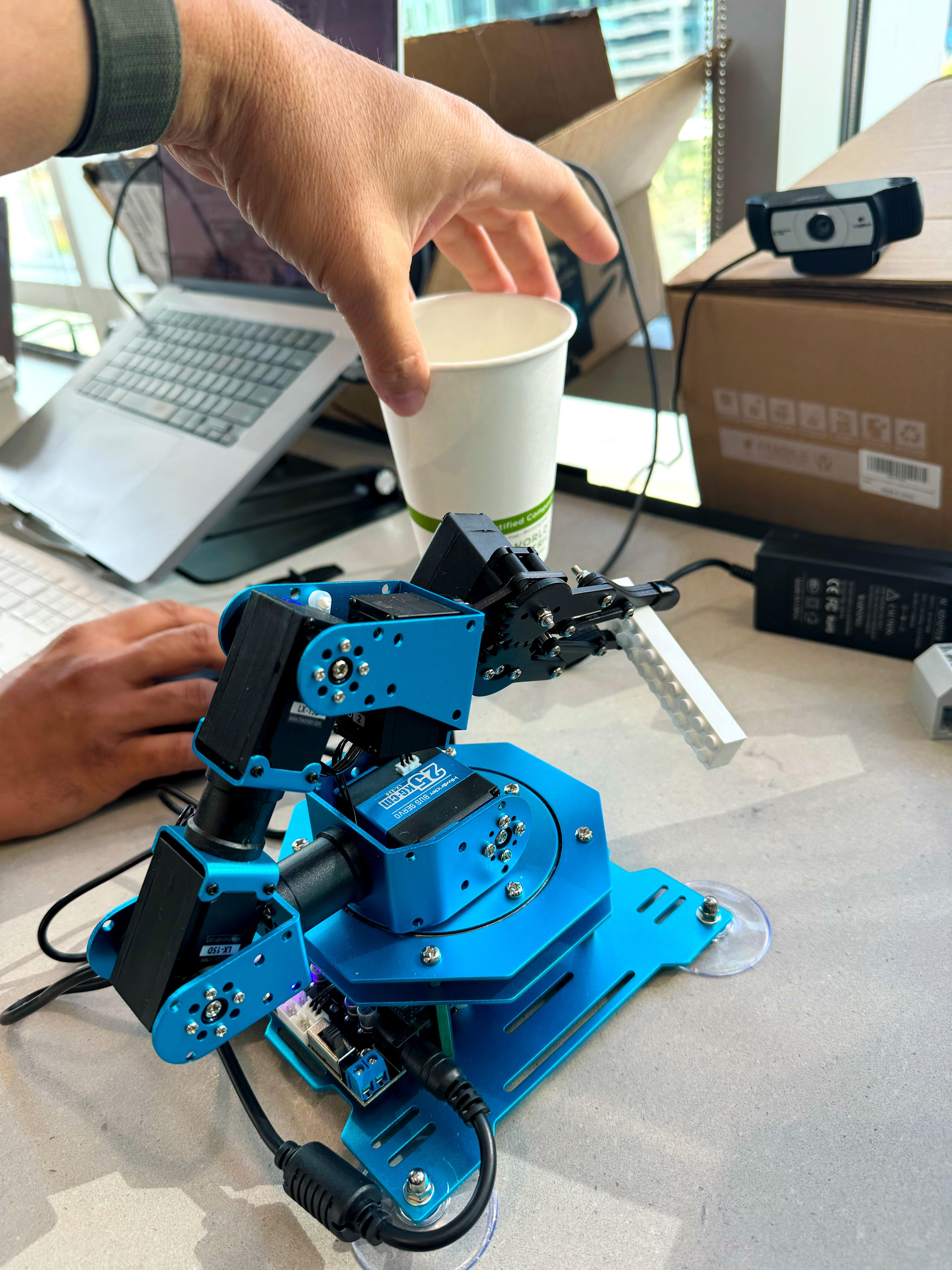 Close-up of the blue robotic arm with a coffee cup and webcam in the background