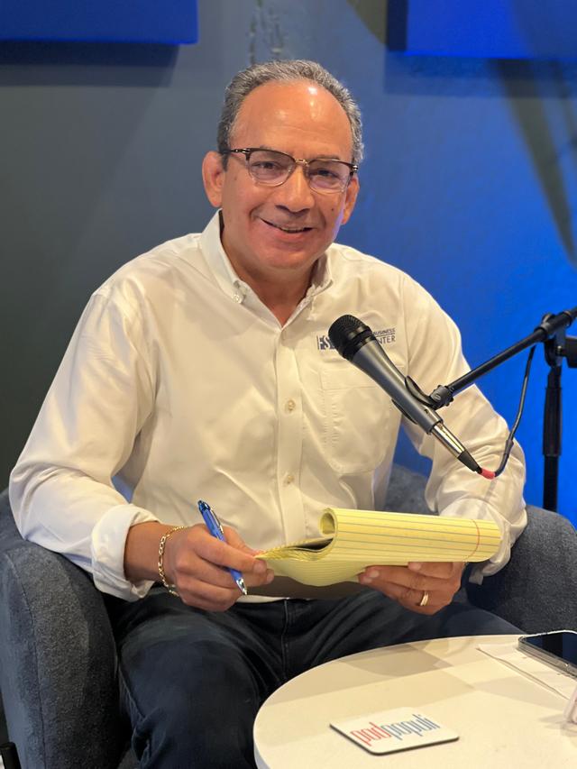 Richard Sierra in a podcast studio setting, wearing a white button-down shirt and glasses, holding a legal pad and pen